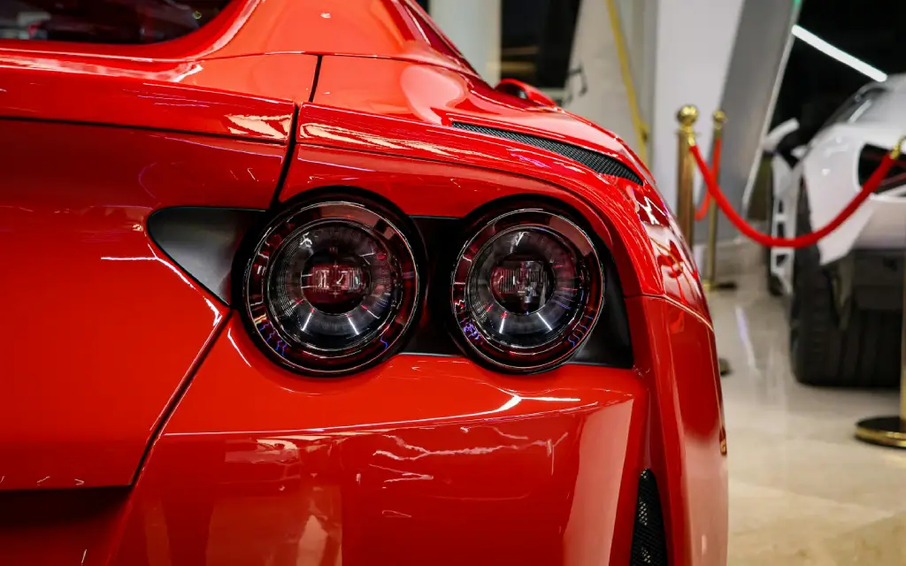 Close-up of the rear left side of a shiny red sports car, focusing on its twin circular taillights, with part of another car and red ropes visible in the background.