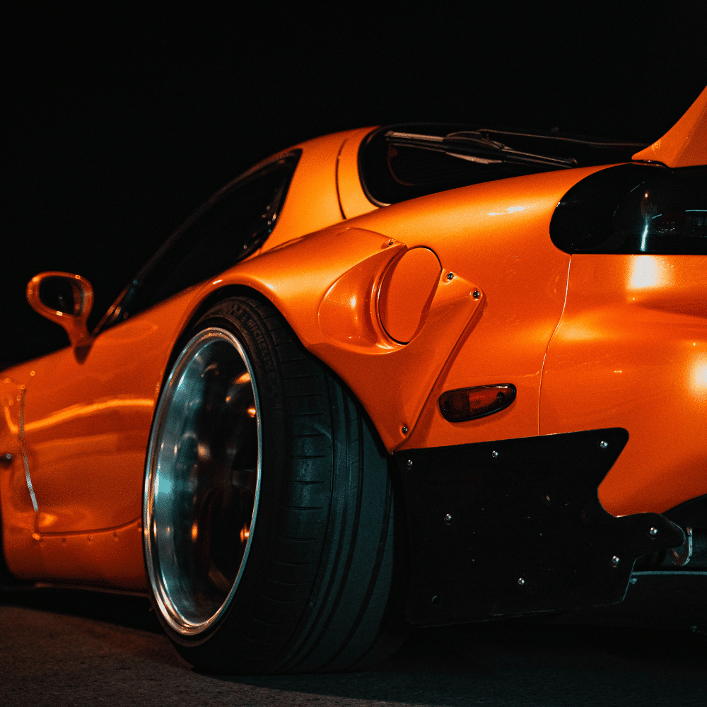 Close-up shot of the rear side of an orange sports car at night, highlighting its wide fender, large tire, and sleek aerodynamic design.