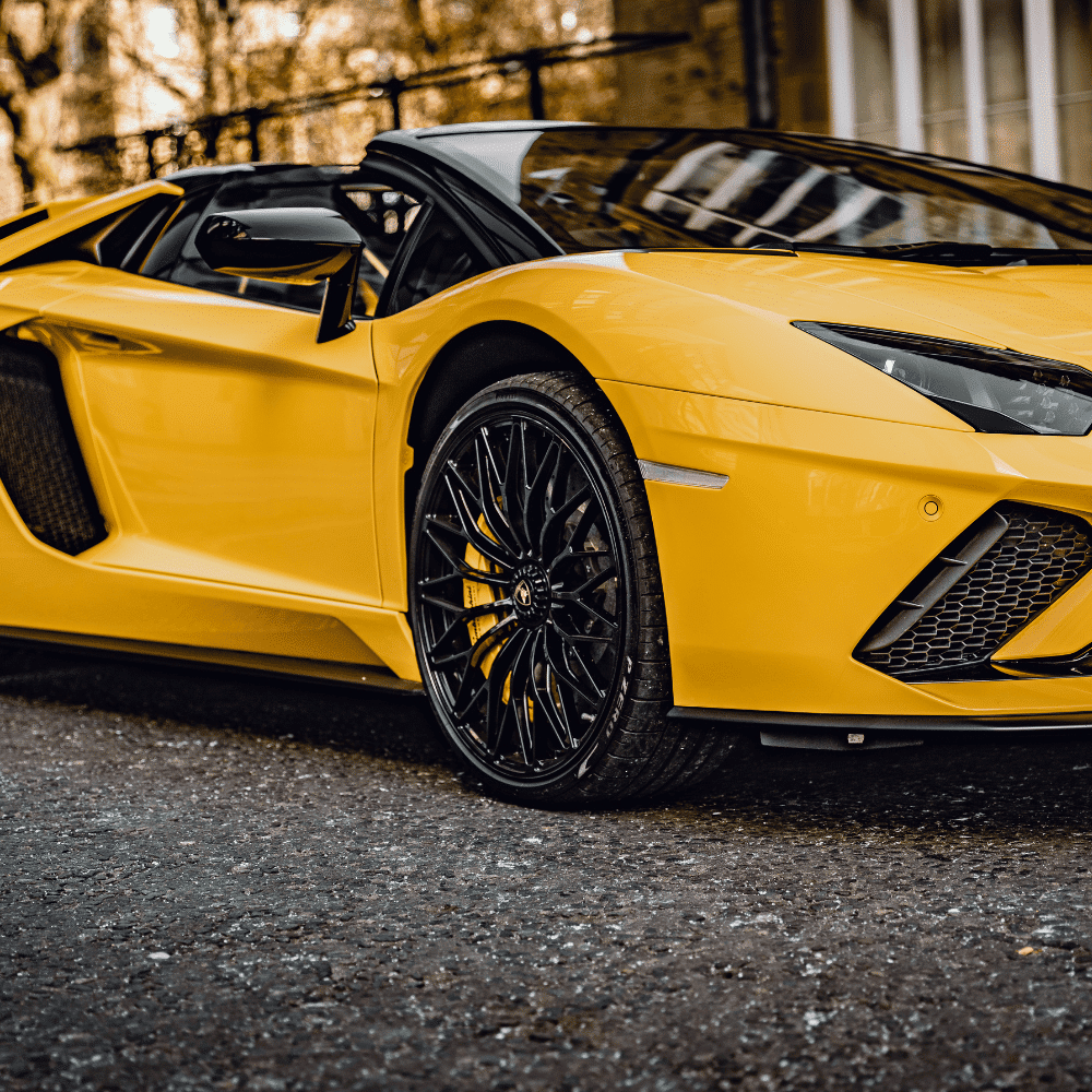 Close-up shot of the rear side of an orange sports car at night, highlighting its wide fender, large tire, and sleek aerodynamic design.