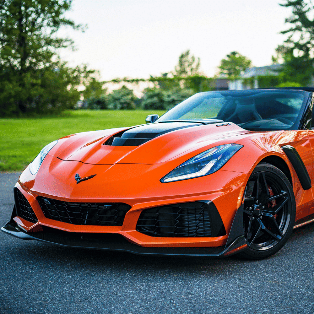 A bright orange Chevrolet Corvette sports car is parked on a paved road with green grass and trees in the background, captured from the front left angle.