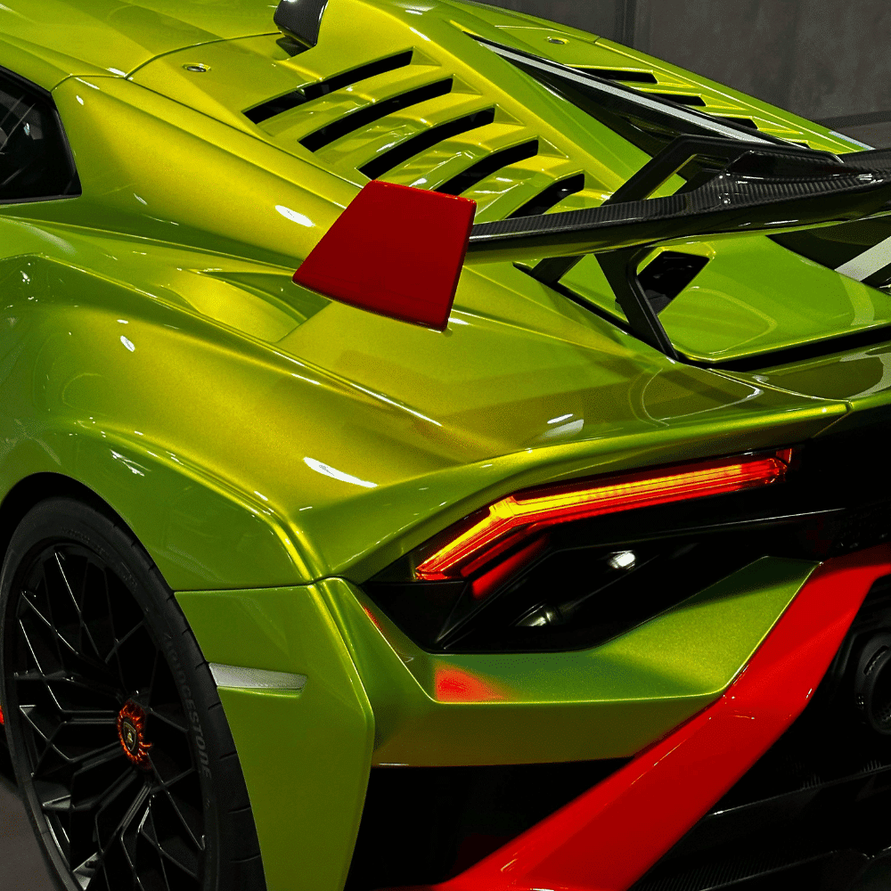 Close-up of the rear section of a bright green sports car with sharp lines, red accents, black rear wing, and angular taillights, showcasing a high-performance, modern automotive design.