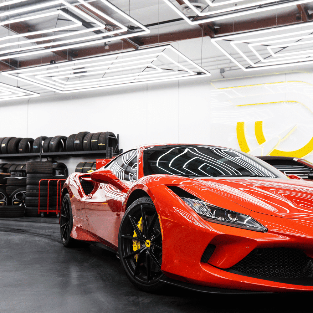A shiny red sports car with black wheels is parked indoors by a wall of stacked tires and modern, geometric ceiling lights reflecting on its surface.