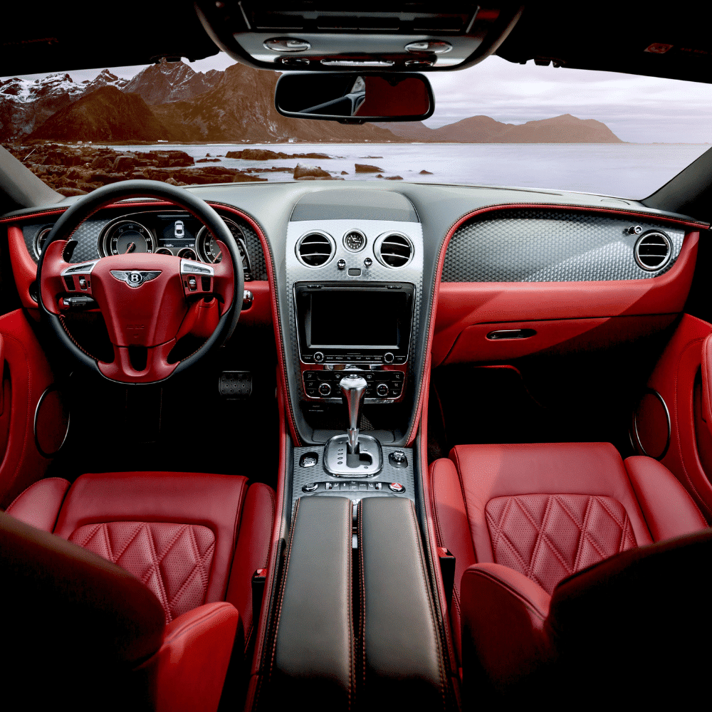 The interior of a luxury car with red leather seats and detailing, a sleek dashboard featuring a large touchscreen, and a scenic ocean and mountain landscape visible through the windshield.