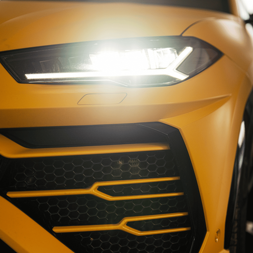 Close-up of the front left headlight and grille of a yellow sports car, with the headlight turned on and a sleek, angular design visible.