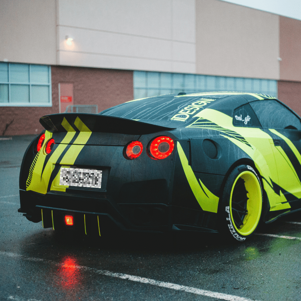 A black sports car with yellow and green geometric decals is parked in a wet, empty lot in front of a brick building. The car has custom rims and a large rear spoiler.